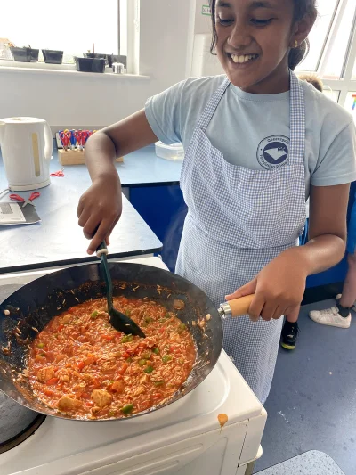 Smiling student in a light blue shirt and blue-checkered apron stirs a tomato-based chicken and veggie sauce in a frying pan on a stove.