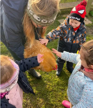 Group of children and an adult pet a brown chicken on a grassy outdoor area, all wearing jackets and knit hats.
