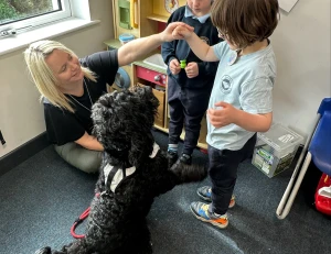 Blonde woman kneels in a classroom by a window as a curly black dog stands on its hind legs, interacting with two children holding a small green toy.
