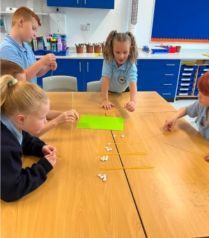 Group of children in a bright classroom building towers with thin sticks and marshmallows on a wooden table, blue cabinets visible in the background.