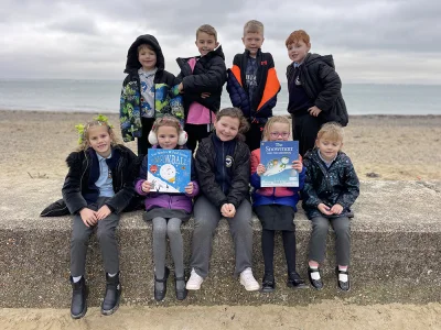 Ten children on a sandy beach, sitting or standing on a concrete wall, wearing jackets; two hold books Snowball and The Snowman and the Snowdog.