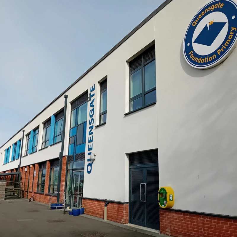 Exterior of a school building with blue windows, white walls and red brick base, featuring a circular Queensgate Foundation Primary sign; blue doors and a yellow wall device.